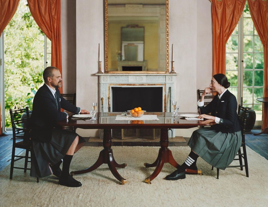 Elegant indoor scene featuring a man and woman seated at a dining table, dressed in formal attire including blazers and pleated skirts.