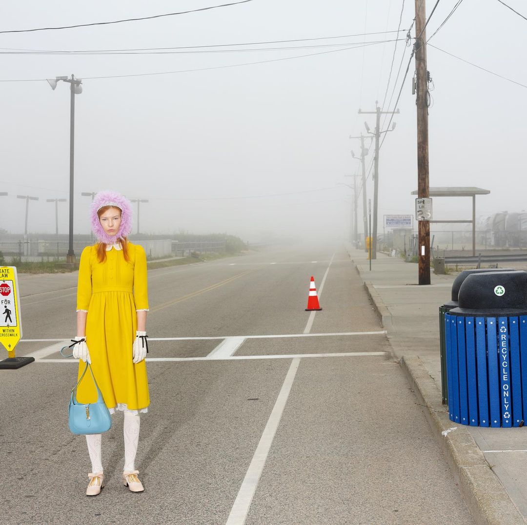 Woman in yellow dress with blue purse on street in dense fog, with a crosswalk sign, trash can, and traffic cone visible.