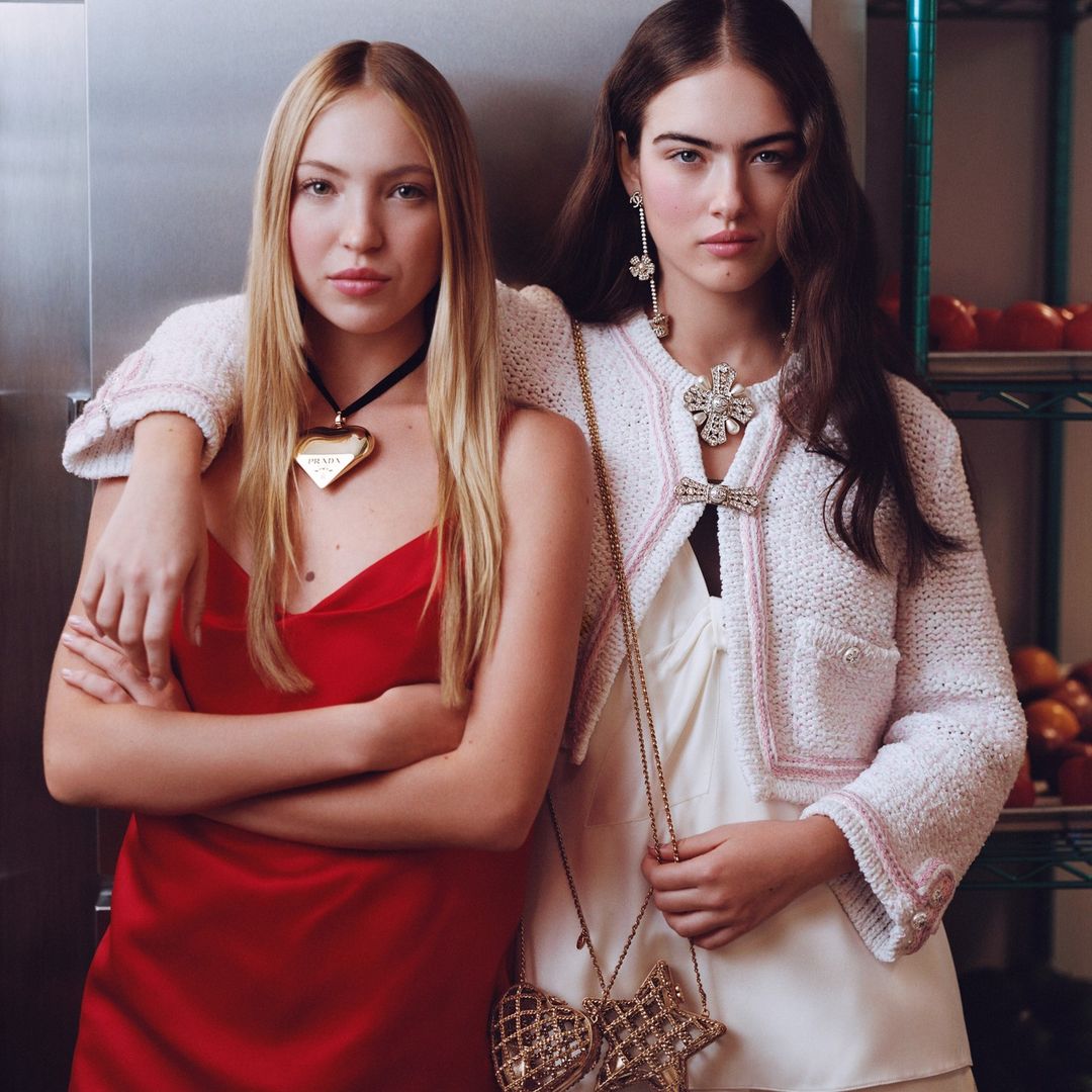 Two women pose indoors; one in red dress with PRADA heart necklace, the other in tweed jacket with chain-strap purse, beauty portrait.