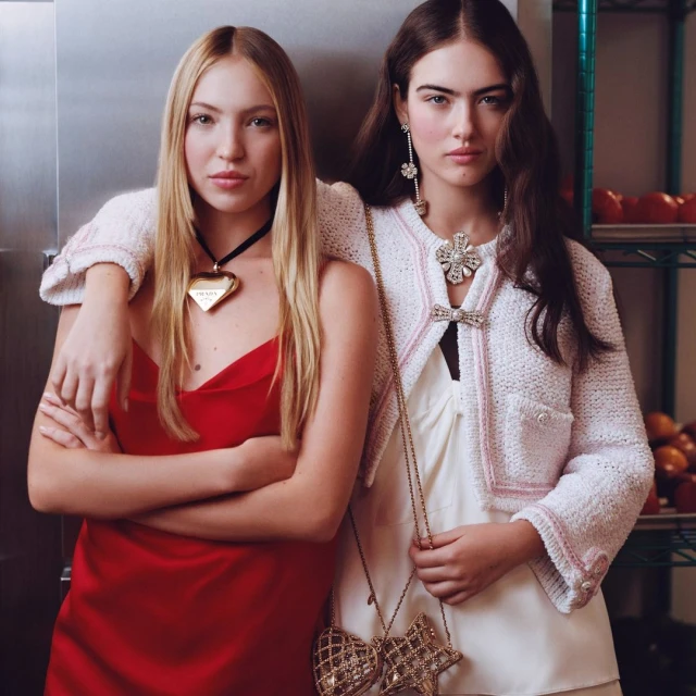 Two women pose indoors; one in red dress with PRADA heart necklace, the other in tweed jacket with chain-strap purse, beauty portrait.