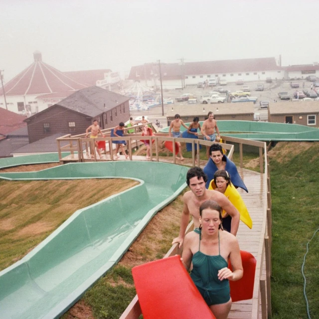 Group of people walking up stairs at a waterpark, carrying colorful mats, with buildings and a parking lot in the background on a cloudy day.