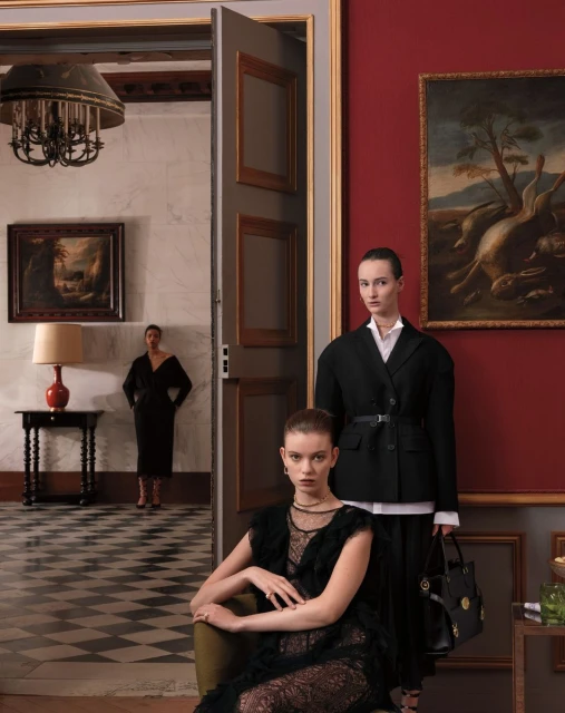 Three women in fashionable black outfits posing indoors near paintings. Interior design features include a checkered floor and ornate chandelier.