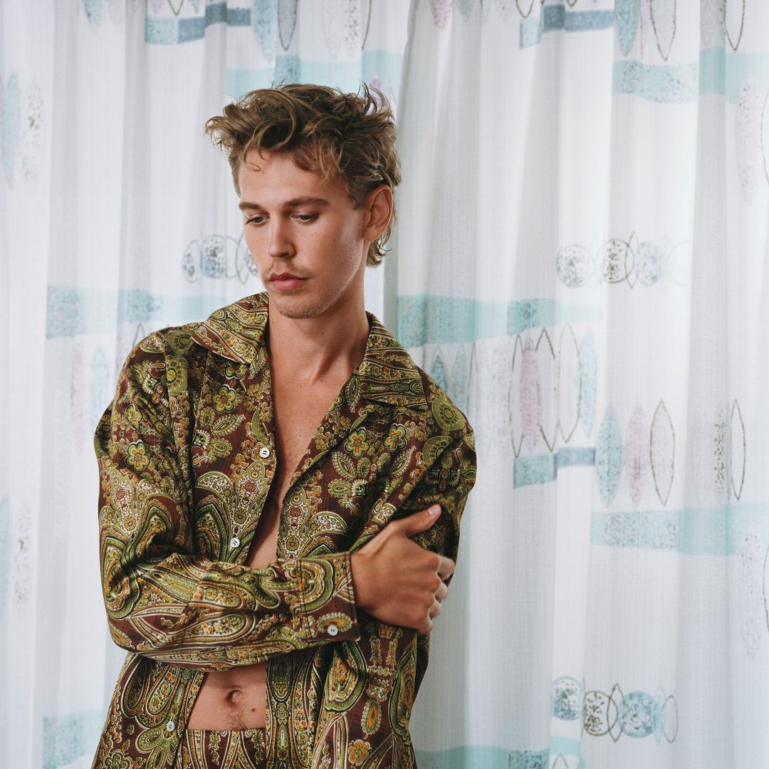 Adult male in patterned shirt posing indoors near patterned curtain, looking down in medium shot with natural lighting. Fashion photography.