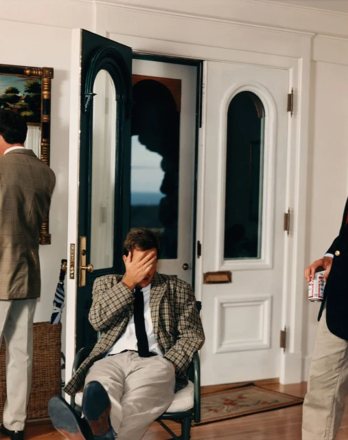Man in plaid blazer sits indoors with his face in his hand while others stand nearby in an indoor scene with white doors and walls.