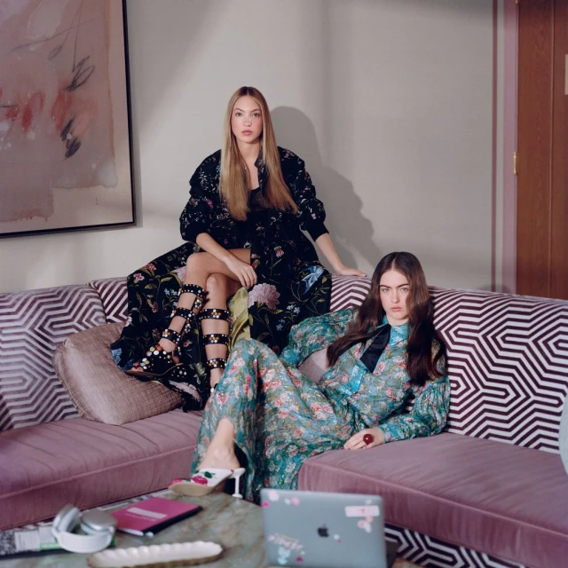 Two women pose on a patterned couch in floral outfits. Laptop, notebook, and dishes on the marble coffee table.
