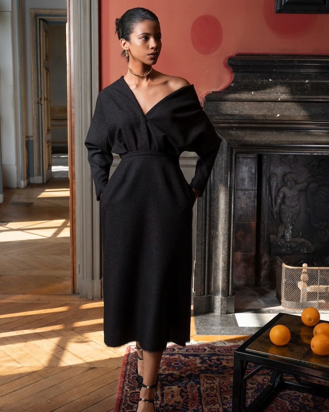 Elegant Black Dress Fashion: A woman showcases a sophisticated black dress indoors, with hardwood floors and a classic fireplace backdrop.