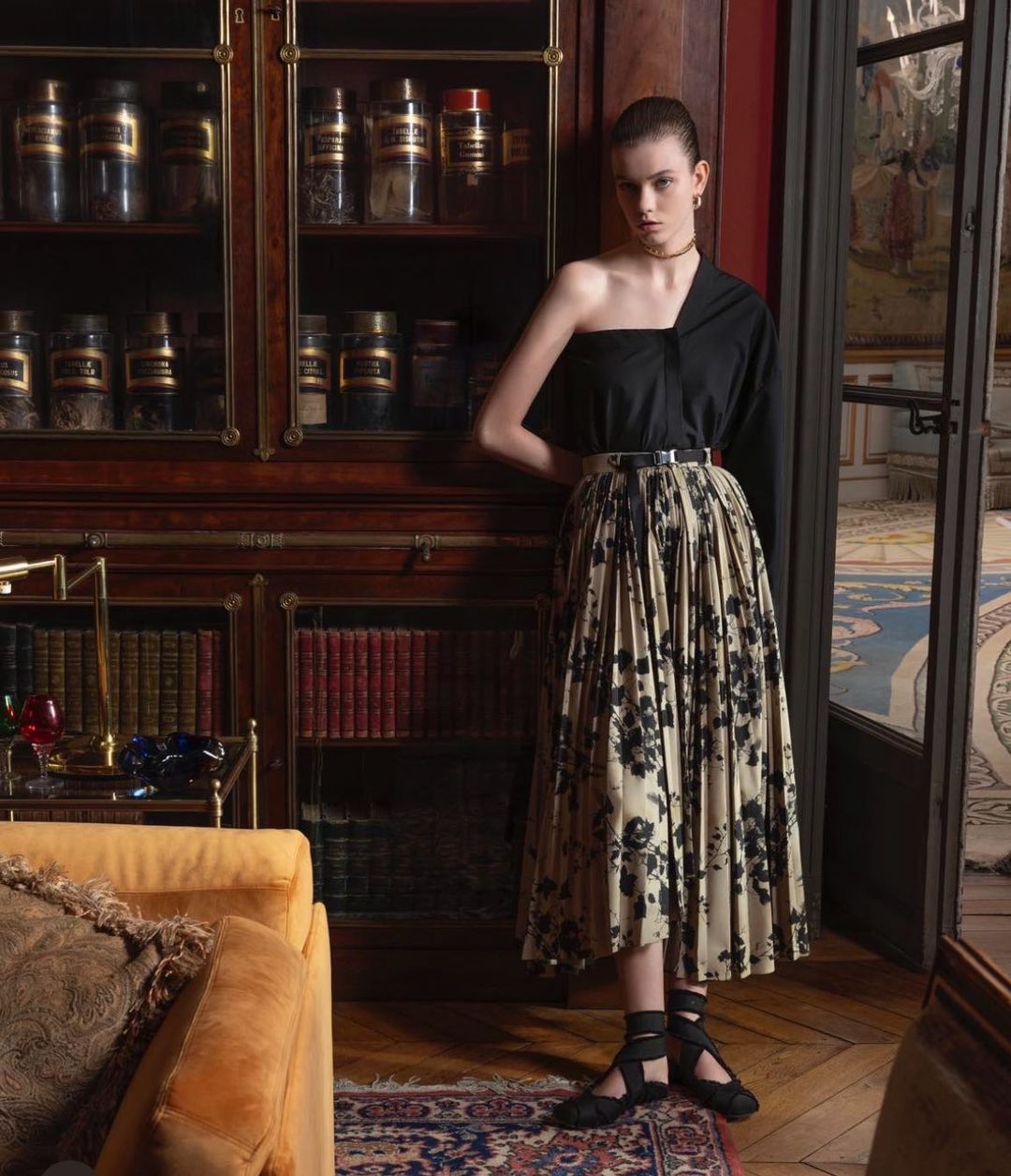 Elegant woman in black one-shoulder top and floral pleated skirt posing indoors near wooden cabinet with glass doors in soft lighting.