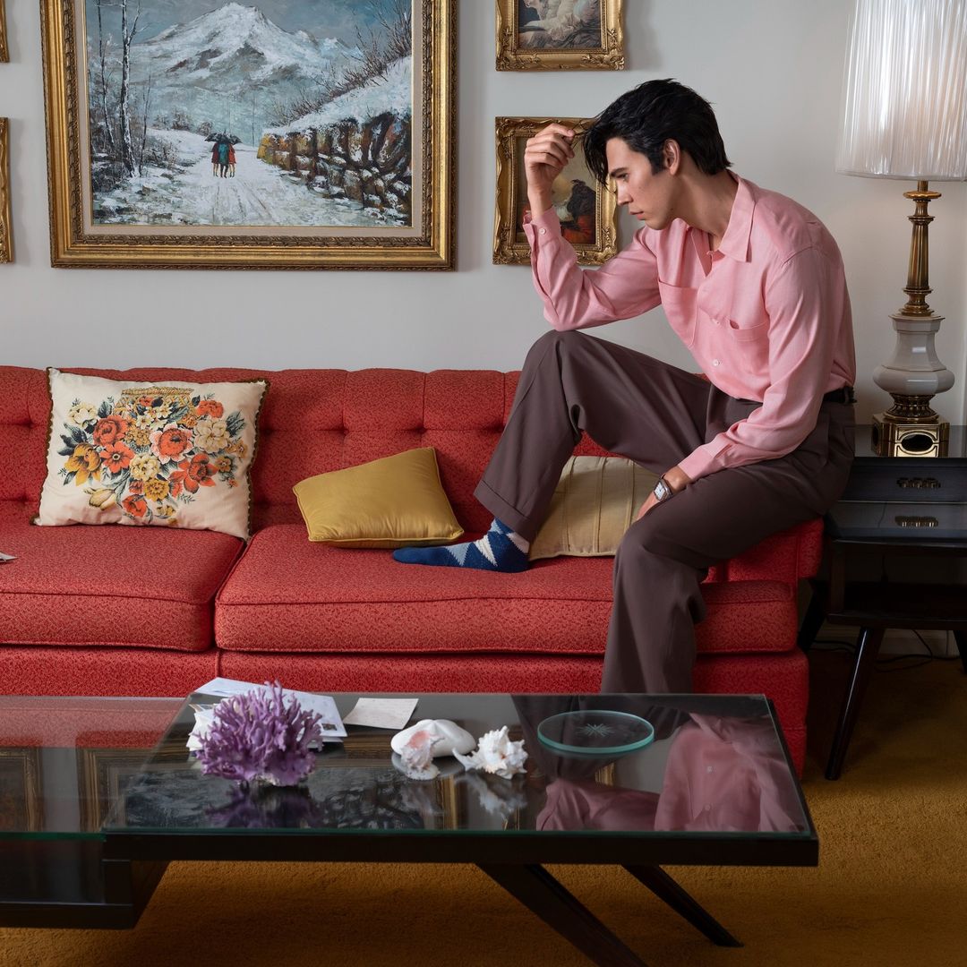 Adult male seated on a red couch in a living room with paintings, glass coffee table, and table lamp, gazing thoughtfully ahead.