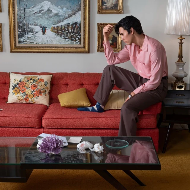 Adult male seated on a red couch in a living room with paintings, glass coffee table, and table lamp, gazing thoughtfully ahead.