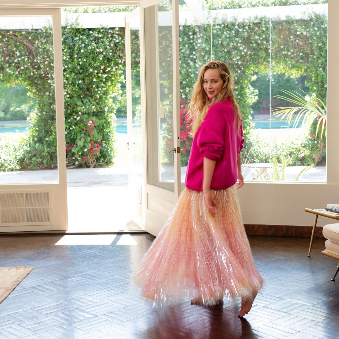 Woman in pink sweater and flowing skirt poses indoors near open doors with lush greenery in the background; natural light, bright.