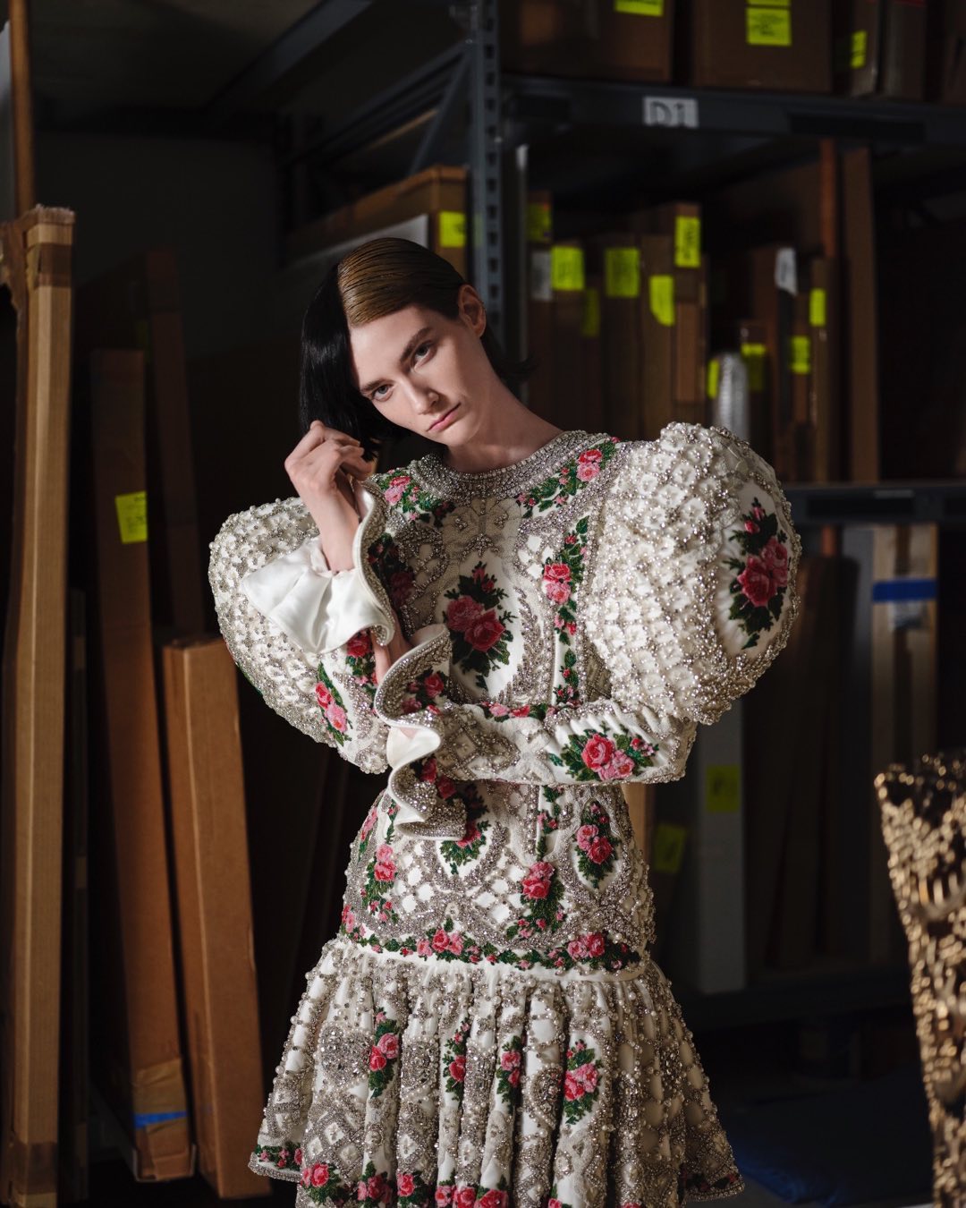 Adult female in a patterned and beaded dress with floral accents, posing in an indoor environment with shelving in the background.