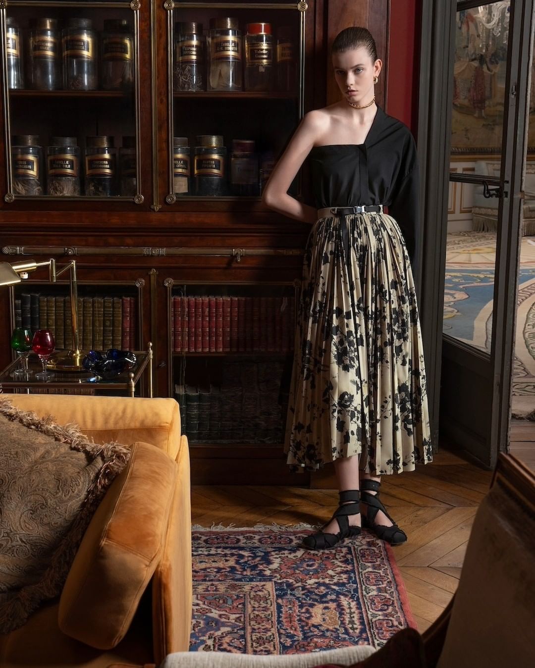 Elegant woman in one-shoulder top and floral pleated skirt posing in a vintage-inspired interior with dark wood furniture and patterned rug.