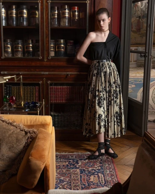 Elegant woman in one-shoulder top and floral pleated skirt posing in a vintage-inspired interior with dark wood furniture and patterned rug.