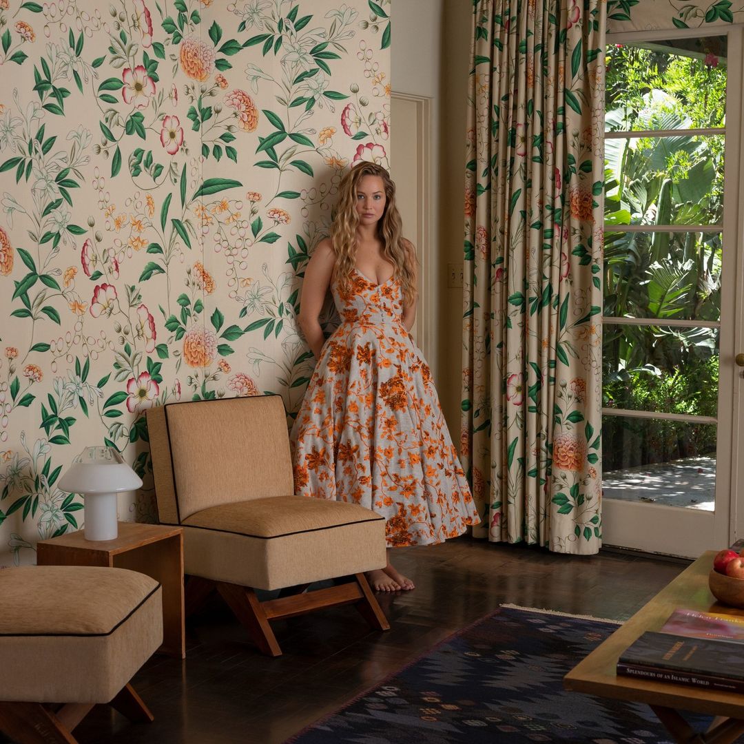 A woman in a floral dress leans against a wall with matching wallpaper, standing in a room with an armchair and view of a lush garden.