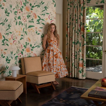 A woman in a floral dress leans against a wall with matching wallpaper, standing in a room with an armchair and view of a lush garden.