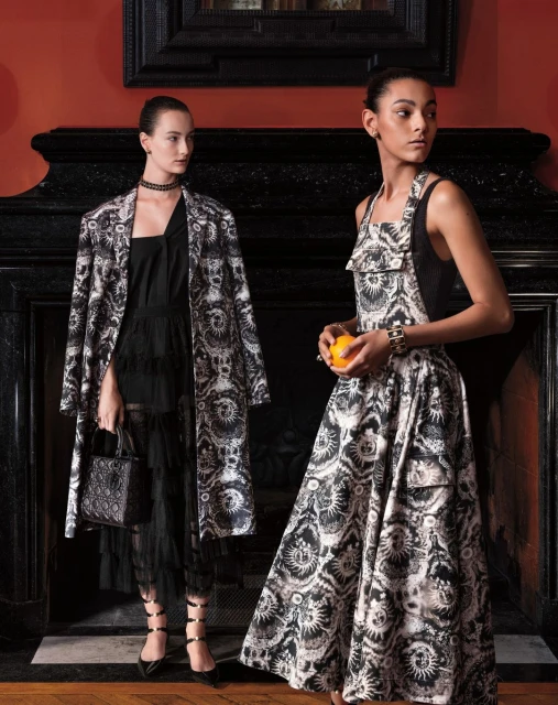 Two fashion models in a black-and-white patterned dress and coat pose in front of a fireplace mantel holding an orange. Interior setting.