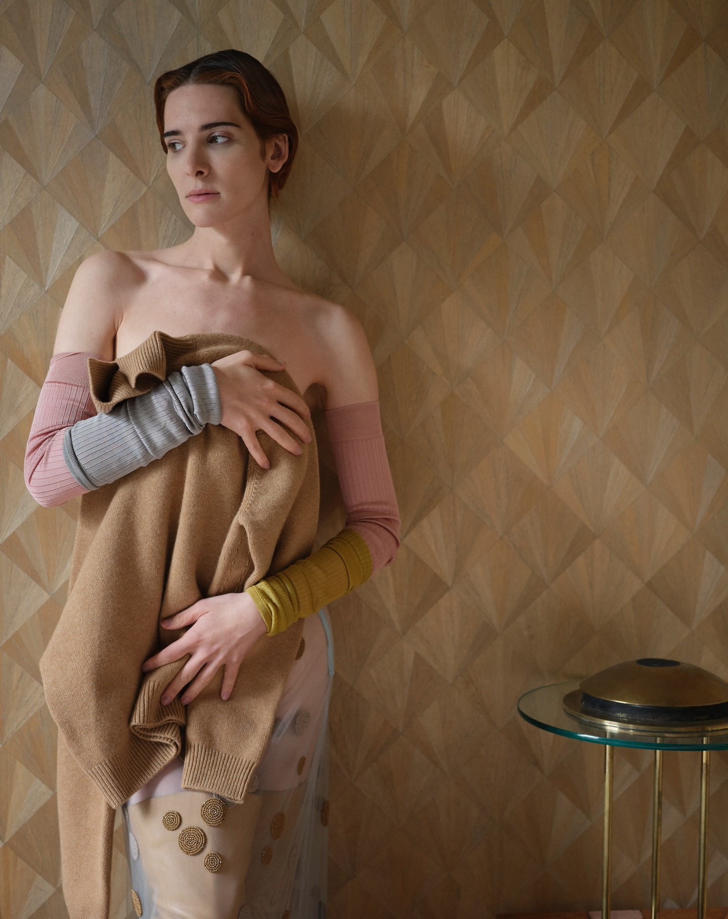 A slender adult woman is indoors, holding a tan sweater in front of her body, wearing long, multi-colored arm coverings.