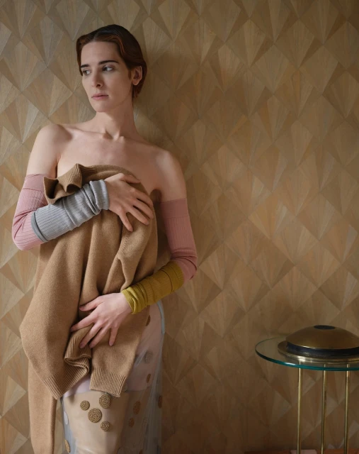 A slender adult woman is indoors, holding a tan sweater in front of her body, wearing long, multi-colored arm coverings.