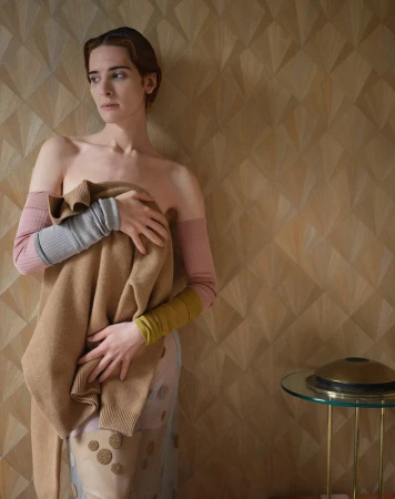 A slender adult woman is indoors, holding a tan sweater in front of her body, wearing long, multi-colored arm coverings.