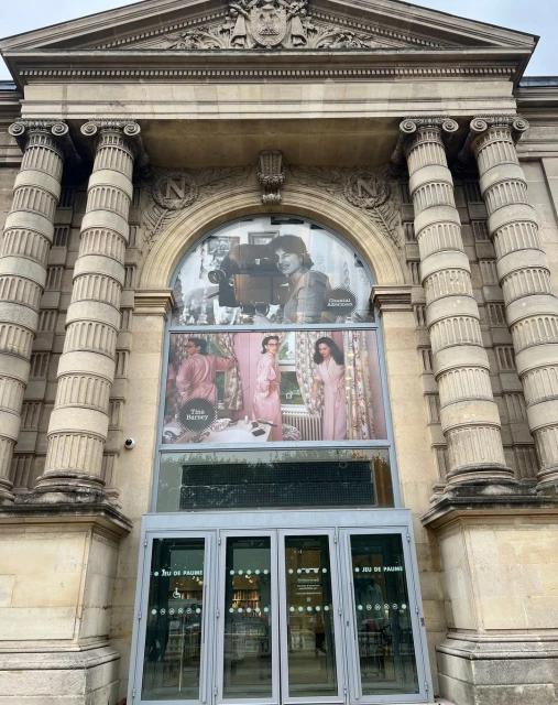 Exterior of Jeu de Paume in Paris featuring a large window with posters of women in pink robes and a vintage camera.