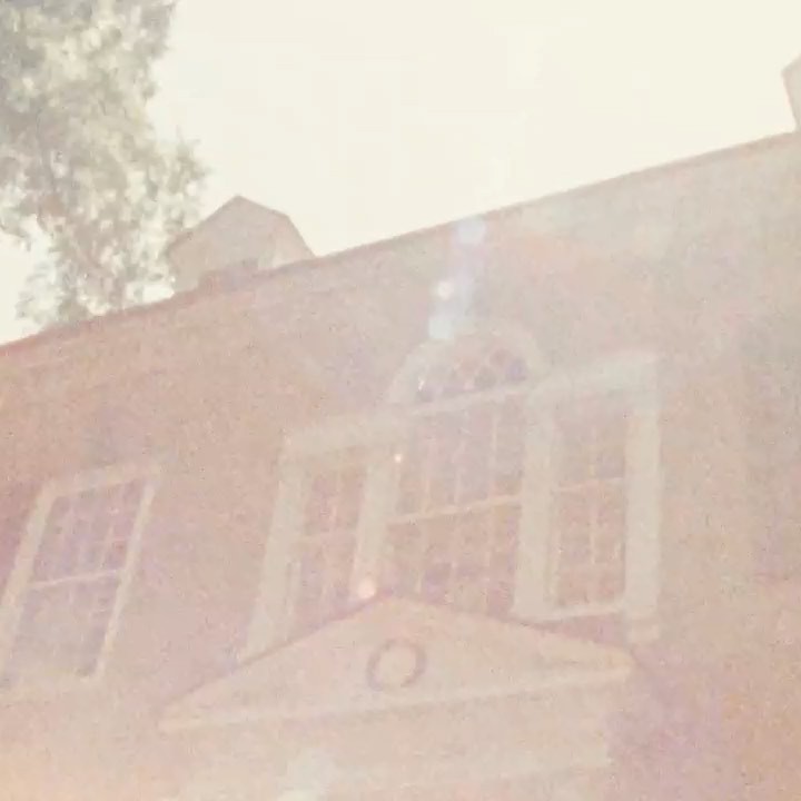 Exterior of a brick building with windows and triangular pediment, shot from a low angle on a bright day. Tree in the background.