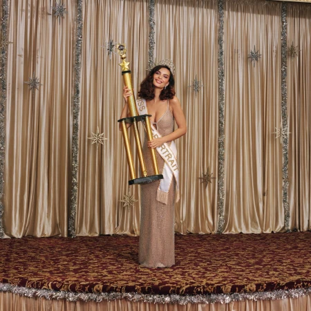 A woman in an evening gown and crown stands on a stage holding a large trophy and a sash that reads "Miss Portrait."