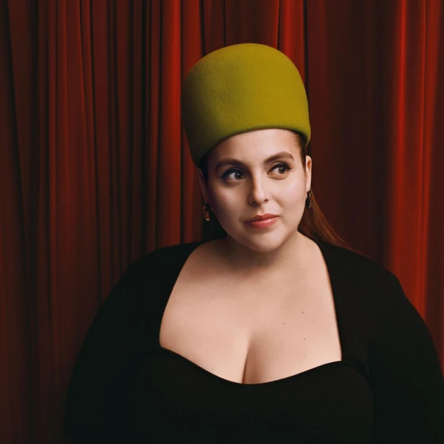Portrait of a woman with fair skin wearing a green hat and a black dress posing in front of a red draped curtain.