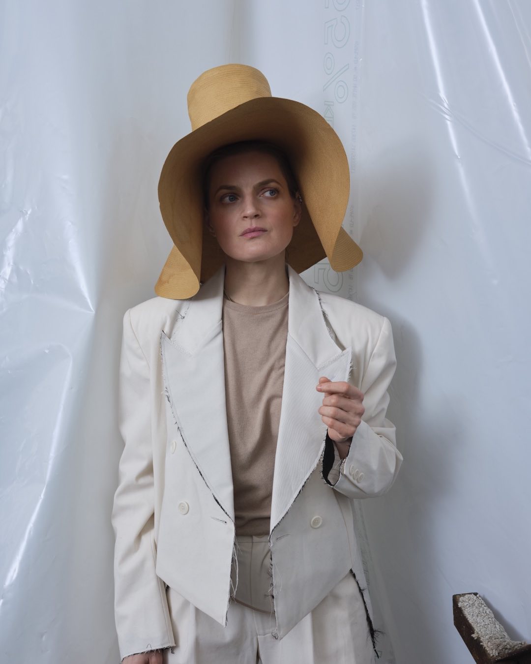 Portrait of a woman wearing a large tan hat and a white jacket against a white background with a partly visible chair in the lower right.