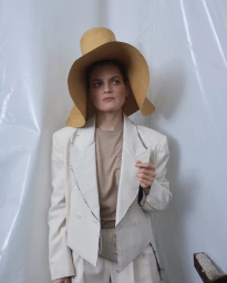 Woman in an oversized white blazer and large yellow hat standing against a textured white plastic sheet backdrop.