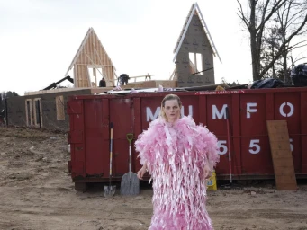 Woman in a textured pink dress standing outdoors in front of a red dumpster and a wooden house under construction.