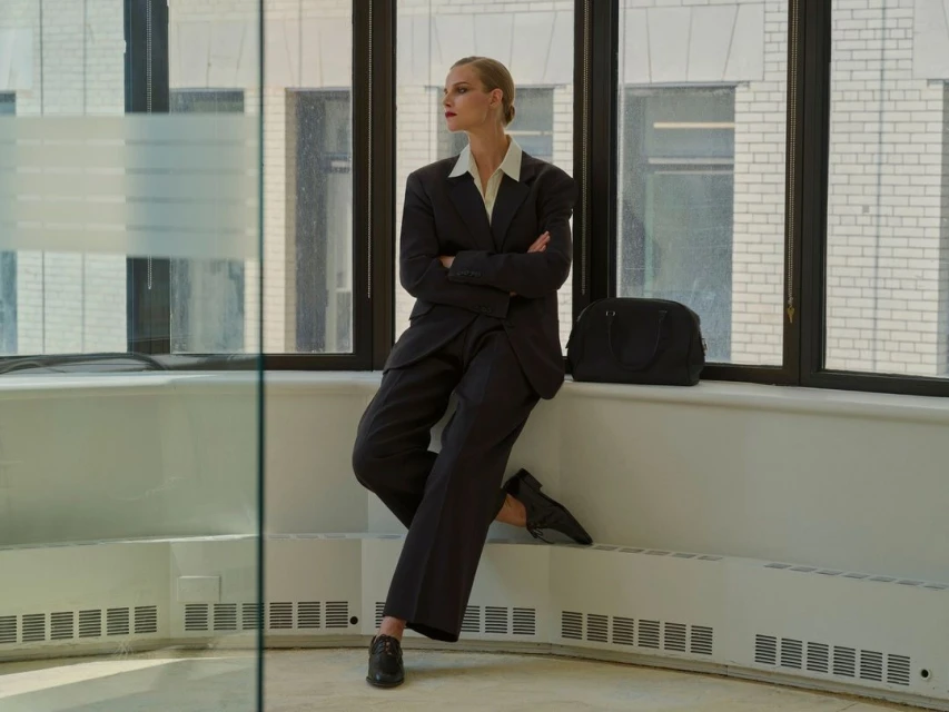 Woman in dark suit poses leaning against a window, indoor office environment, wearing dark shoes and standing near a black duffel bag.