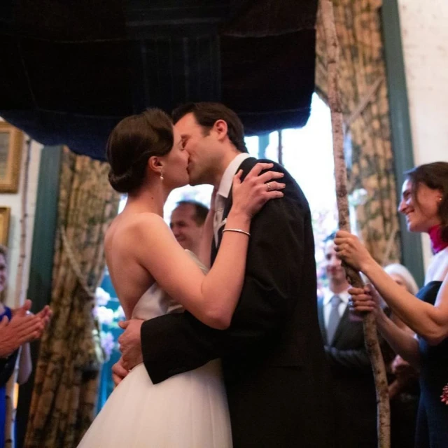 Bride and groom kiss at their wedding ceremony indoors under a chuppah while guests look on and applaud.