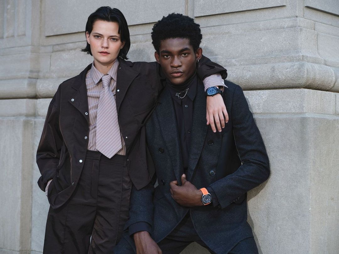 Two people in business attire pose outdoors. The man has his arm on the woman's shoulder. They are both wearing wristwatches.