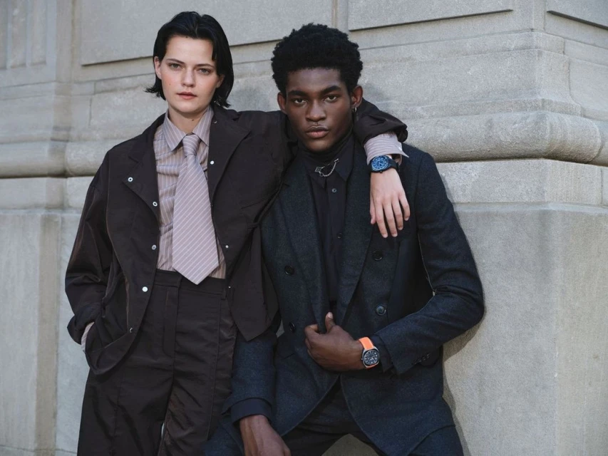 Two people in business attire pose outdoors. The man has his arm on the woman's shoulder. They are both wearing wristwatches.