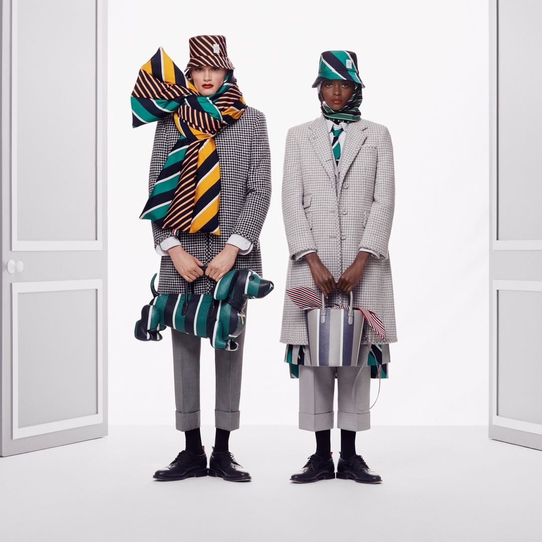 Two fashion models in houndstooth coats, hats, and scarves, standing in a white photo studio with a dog-shaped purse and striped tote bag.