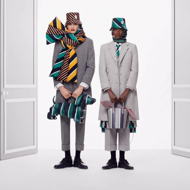 Two fashion models in houndstooth coats, hats, and scarves, standing in a white photo studio with a dog-shaped purse and striped tote bag.