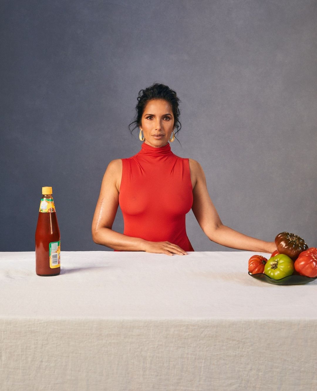 A woman in a red turtleneck poses at a table with various tomatoes and a bottle of hot sauce against a gray background.