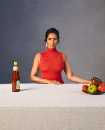 A woman in a red turtleneck poses at a table with various tomatoes and a bottle of hot sauce against a gray background.