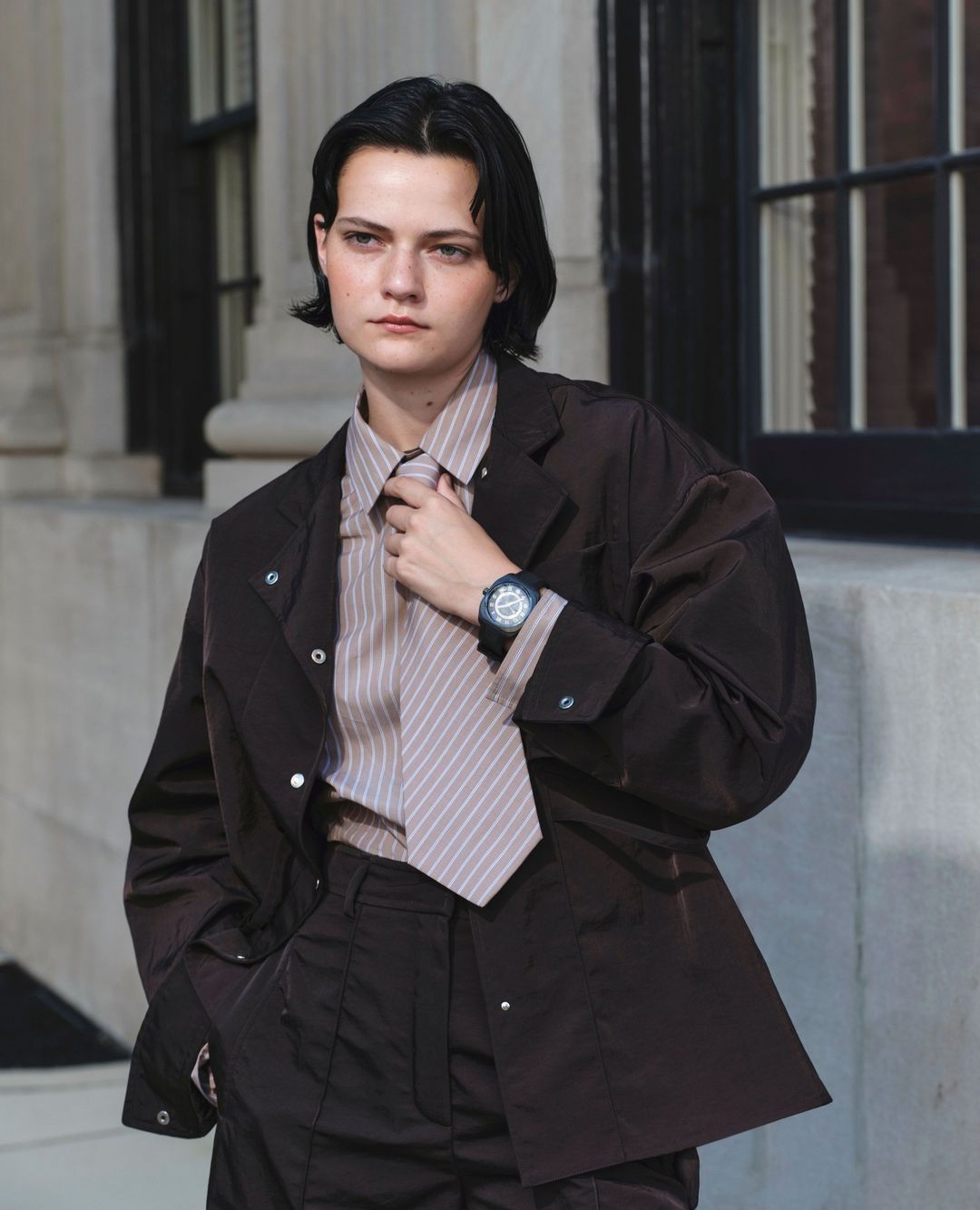 Adult female model in a brown jacket, pants, and striped shirt posing outdoors, looking at the camera with neutral expression.