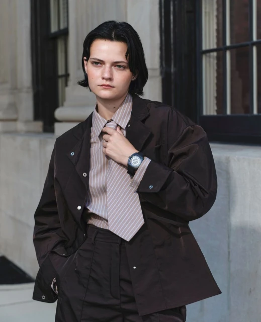 Adult female model in a brown jacket, pants, and striped shirt posing outdoors, looking at the camera with neutral expression.