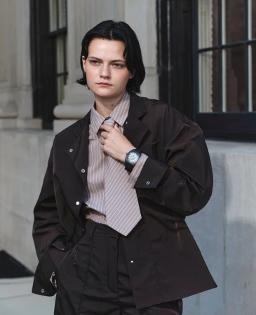 Adult female model in a brown jacket, pants, and striped shirt posing outdoors, looking at the camera with neutral expression.