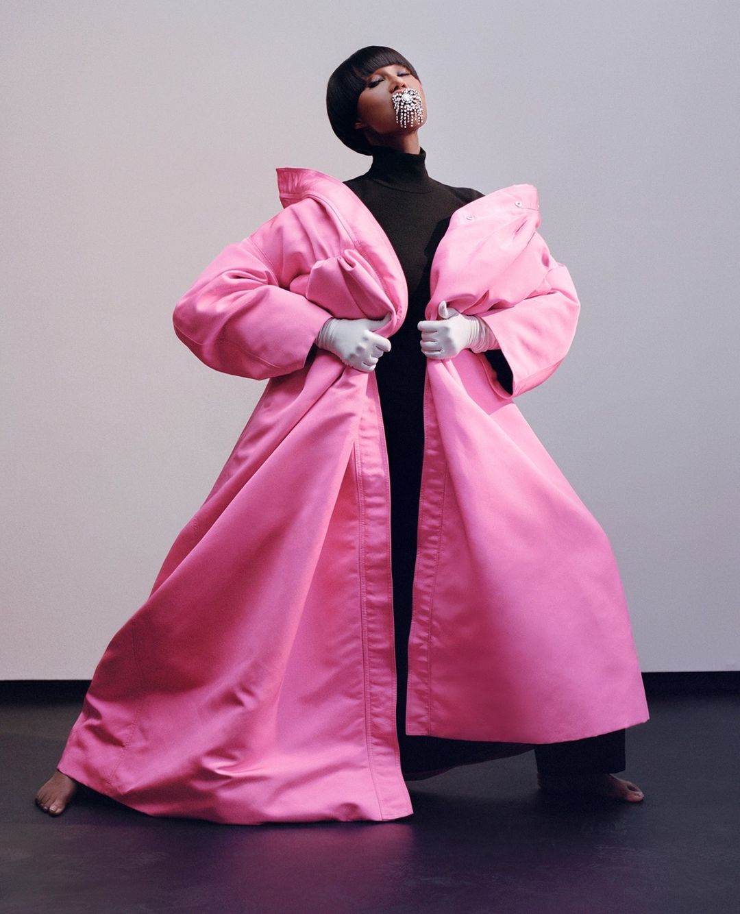 A woman in a large pink coat with a black turtleneck and wide-leg black pants strikes a pose in a photo studio with a grey backdrop.