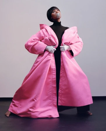 A woman in a large pink coat with a black turtleneck and wide-leg black pants strikes a pose in a photo studio with a grey backdrop.