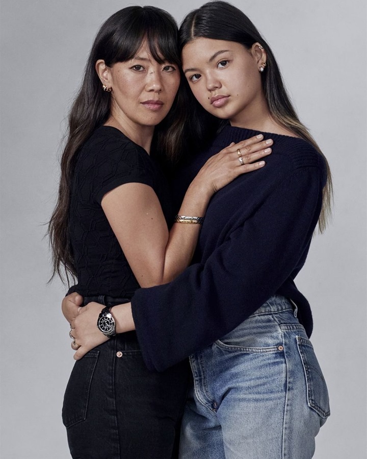 Two Asian women embrace in a studio portrait. The woman on the left wears a black top and jeans, while the other wears a blue sweater and jeans.