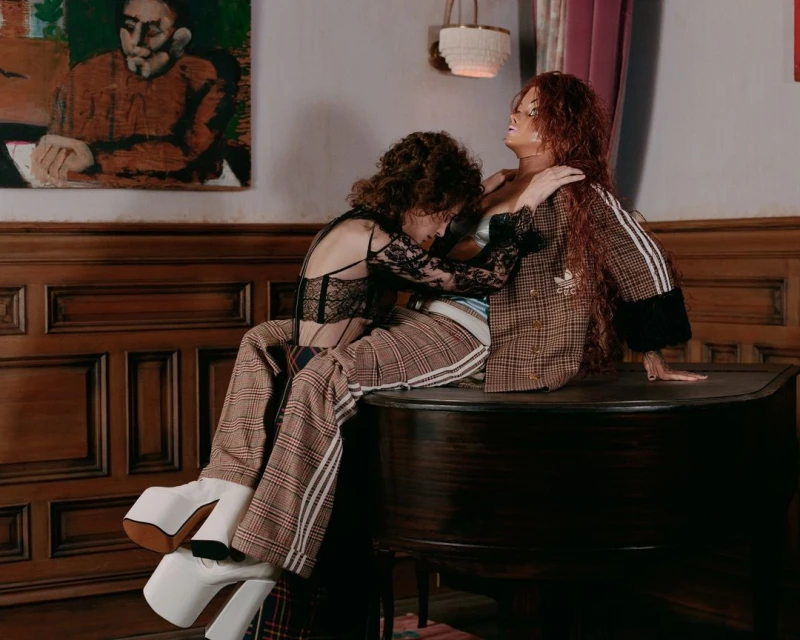 Two models pose in an indoor setting. One model sits on the piano while the other leans in, both wearing fashionable clothing.
