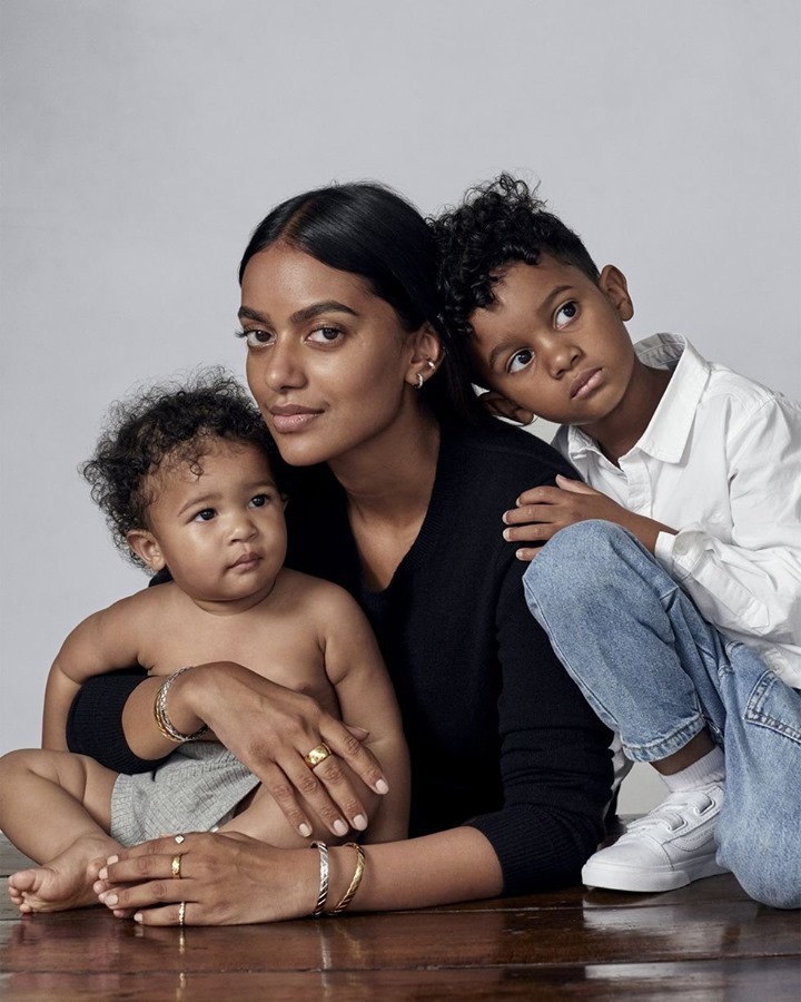 Portrait of a dark-haired woman with two children. The infant sits on her lap, while the older child leans in from behind. All are wearing jewelry.