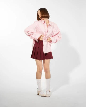 A young woman in a pink button-down shirt and a maroon skirt poses against a white background in a fashion photography style.