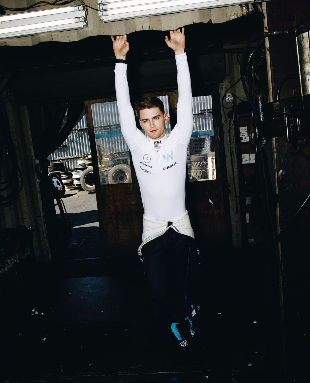 A man in a racing suit hangs from a garage ceiling, wearing a white racing suit, with auto tires in the background and fluorescent lighting.