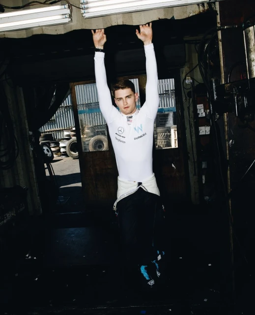 A man in a racing suit hangs from a garage ceiling, wearing a white racing suit, with auto tires in the background and fluorescent lighting.