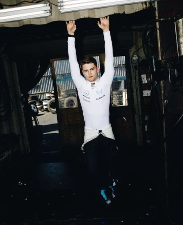 A man in a racing suit hangs from a garage ceiling, wearing a white racing suit, with auto tires in the background and fluorescent lighting.